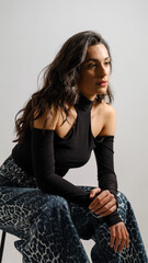 In a studio setting with a white background, a young black-haired woman wearing a black blouse and jeans strikes an artistic pose