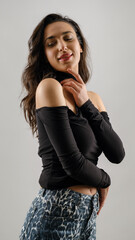In a studio setting with a white background, a young black-haired woman wearing a black blouse and jeans strikes an artistic pose