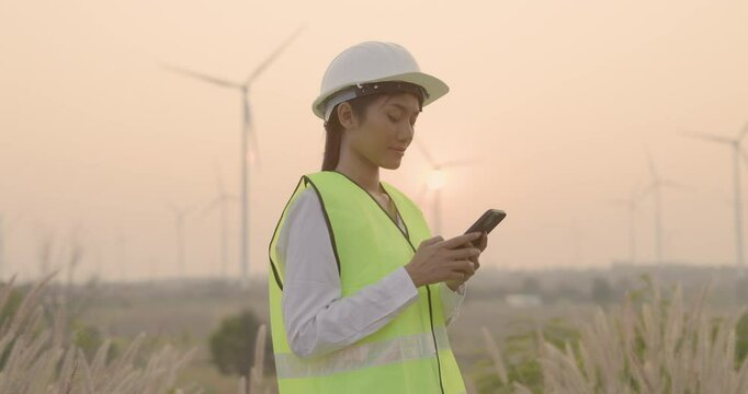 Portrait of female worker or asian woman engineer working with mobile phone at wind turbine farm. Wind power turbines generating clean renewable energy for sustainable development.