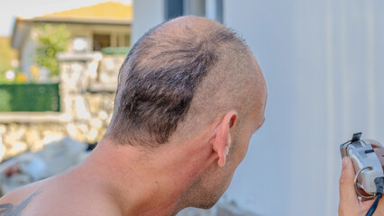 Fototapeta premium A handsome middle-aged man shaving his hair and beard topless in the tree-filled garden of his house on a sunny summer day
