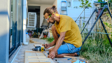 A middle-aged man measures with a tape measure to make repairs with lumber in his garden on a...