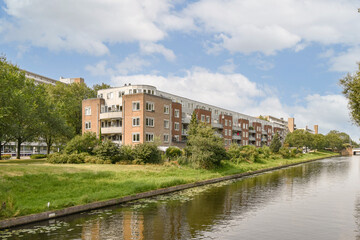 Fototapeta premium A serene view of a river lined with modern residential buildings and lush greenery under a blue sky. Ideal for showcasing urban life and nature.