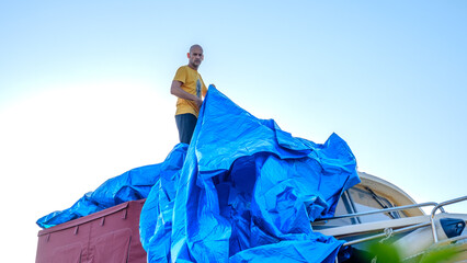 A middle-aged man covering a boat on land with a blue tarp in the garden of his house on a summer day