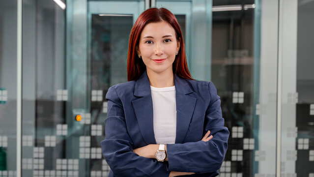 A young, fair-skinned businesswoman with red hair stands in an empty office environment with a blurred background, arms crossed, smiling as she looks at the camera