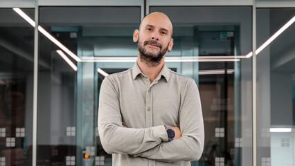 Obraz premium A middle-aged, fair-skinned businessman is in an empty office environment with a blurred background, crossing his arms and smiling as he looks at the camera