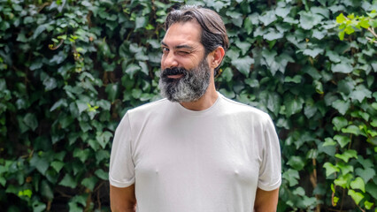 A middle-aged, fair-skinned, dark-haired man with a beard is outdoors in a wooded area, winking at...