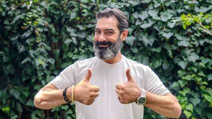 A middle-aged, fair-skinned, bearded dark man outdoors in a wooded area, looking at the camera and giving a thumbs-up