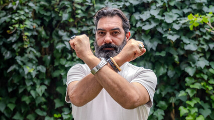 A middle-aged man with fair skin and a beard, outdoors in a wooded area, making a "no" gesture with his hands by crossing them, while looking at the camera