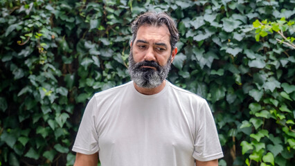 A middle-aged, light-skinned, bearded, dark-haired man looking at the camera with a sad expression in a wooded outdoor setting