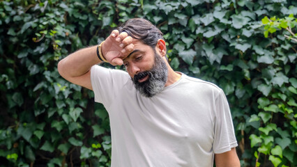 A middle-aged, dark-haired man with a beard and fair skin, outdoors in a wooded area, looking at the camera in close-up with an expression showing discomfort from the heat