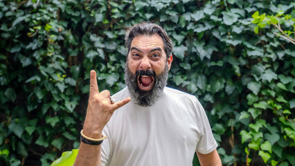 A middle-aged, fair-skinned, bearded, dark-haired man making the metal music hand gesture while looking at the camera in a wooded outdoor setting
