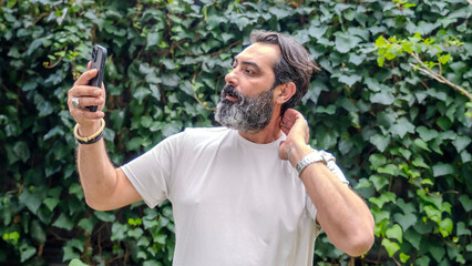 A middle-aged, fair-skinned, bearded, dark-haired man taking a selfie with his cellphone in a wooded area, in a close-up shot