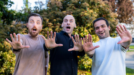 Three middle-aged men pose in surprise in a wooded area, their smiles and gestures radiating joy...