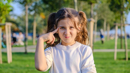 Naklejka premium A little girl standing in a wooded park on a spring day, looking at the camera while placing her hand on her forehead, showing a thoughtful expression