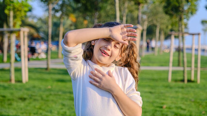 Obraz premium A little girl child standing in a wooded park on a fall day, looking at the camera and making facial expressions that show she is sweating