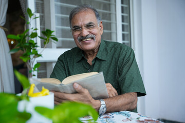 Happy Senior indian man with eyeglasses reading book as hobby at home, Mature elderly male...