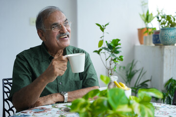 Happy retired senior indian man in 60s drinking morning tea or coffee sitting in balcony, Mature...