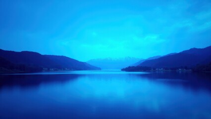 Obraz premium A serene lake surrounded by mountains under a blue hue sky.