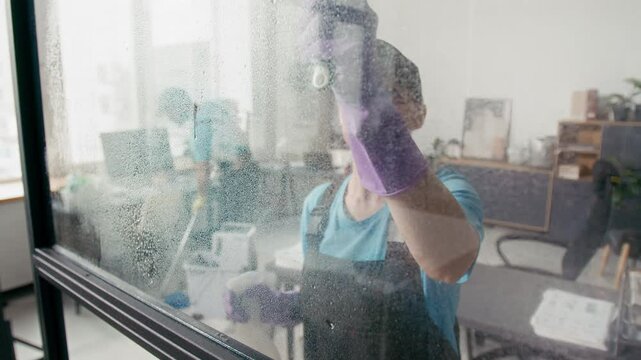 Caucasian woman in uniform and protective gloves disinfecting surface of glass wall while male colleague mopping floor in background at office