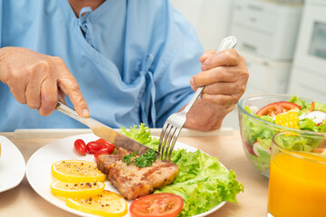 Asian senior woman patient eating pork chop stake and vegetable salad for healthy food in hospital.