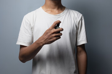 A man wearing a white t-shirt holds a bottle of perfume and sprays it on his body area.