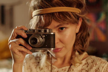 Portrait of young woman holding vintage camera, wearing headband, and looking intently at the lens,...