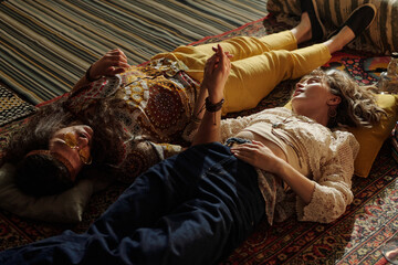 Two people lying on a patterned carpet, resting with closed eyes in a relaxed atmosphere. One...