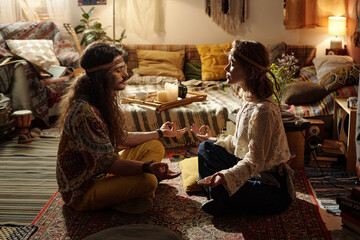 Two people meditating in a cozy, bohemian-style living room with various decorations. Sitting on a...