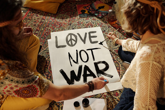 Group of people painting an anti-war poster with brushes and inks. Colorful patterned rug providing backdrop, artwork conveying peace message written in bold letters - Powered by Adobe
