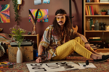 Long-haired man sitting on floor surrounded by colorful artwork and plants while making a poster....