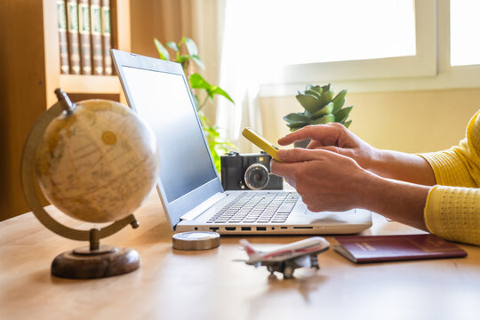 Woman using smartphone and laptop booking a trip online with globe, passport, plane model, compass and camera on a wooden table in front of bright window