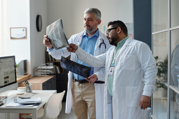 Obraz premium Side view portrait of two mature doctors holding X-ray images and discussing medical case results while standing in clinic office copy space