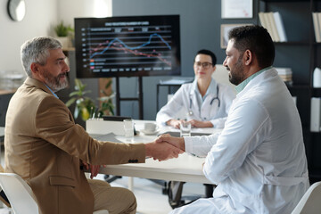 Fototapeta premium Side view portrait of mature businessman shaking hands with smiling Middle Eastern doctor during medical conference copy space