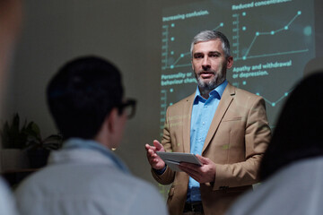Waist up portrait of mature businessman speaking to audience at medical conference and holding digital tablet while explaining statistics data copy space