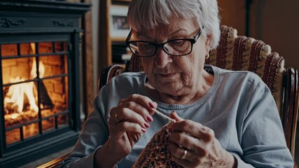An elderly woman is focused on knitting in a warm living room. The fireplace burns brightly, creating a cozy atmosphere as she enjoys her craft on a winter day. - Powered by Adobe