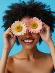 Horizontal shot of pretty woman has Afro hair holds two gerbera daisy in front of eyes and smiles broadly, has fun. Fashionable lady enjoys spring time, pleasant fragrance. Florist with flowers