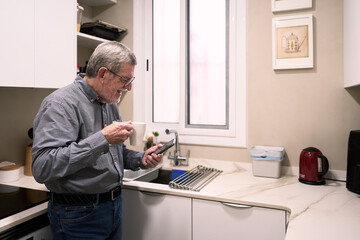 Senior man enjoying coffee and using smartphone in modern kitchen