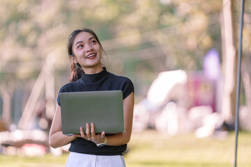 Happy young asian woman freelancing in a park, holding a laptop and smiling while looking up. Enjoying remote work amidst nature, surrounded by greenery and sunlight