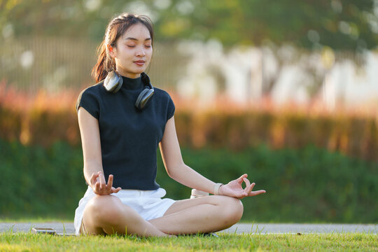 Serene young asian woman finds tranquility as she practices yoga and meditation outdoors in a park, wearing headphones and sitting in the lotus position on the grass
