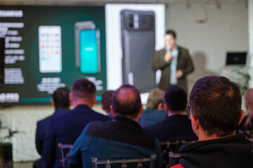 Audience listening to speaker during business presentation featuring latest smartphone technology with display screens