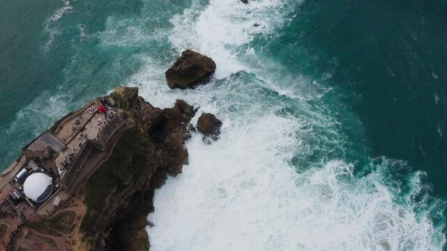 Farol da Nazar&eacute; - cliffs with lighthouse washed by Atlantic ocean waves crashing over rocky coast during international surfers challenge in Nazare, Portugal on February day. 4K aerial top view video