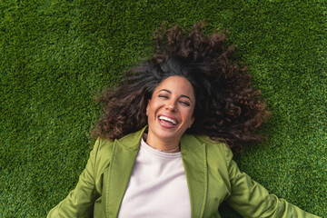 Happy Woman Lying on Grass Laughing. Joyful woman with curly hair lying on green grass, smiling and laughing. Concept of happiness, relaxation, and positivity.