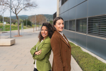 Fototapeta premium Confident Businesswomen Standing Back to Back Outdoors. Two professional women in green and brown blazers smiling while standing back to back near a modern office building