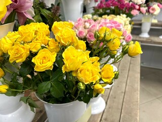 Close-up of yellow roses standing in a white vase on a background of various flowers on a wooden table in a flower shop to make a bouquet with home delivery