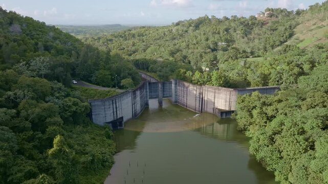 Aerial approaching shot of Hatillo Dam with wall and water at sunny day. Green forest landscape in Sanchez Ramirez, Dominican Republic.