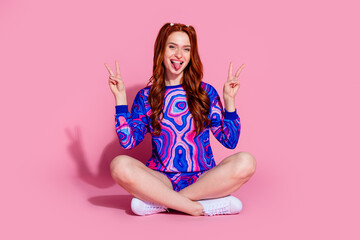 Full body photo of attractive young woman sit floor v-sign stick tongue out dressed blue print clothes isolated on pink color background