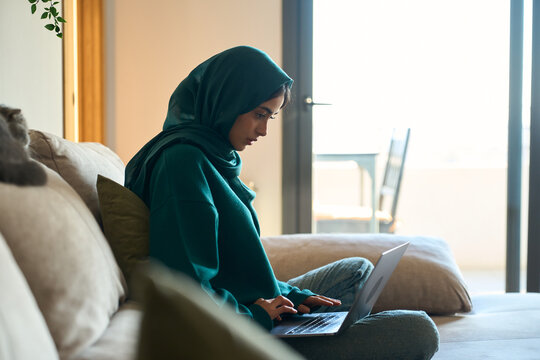 Young muslim woman wearing hijab working from home using laptop on sofa