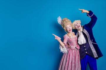Elegant couple in extravagant baroque attire joyfully pointing in front of a vibrant blue backdrop