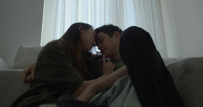 A couple sits closely on a couch, sharing tender gestures and gazes.