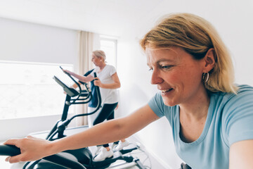 Middle-aged women exercising on elliptical machines - Smiling women working out on elliptical trainers in a bright, modern gym, promoting active and healthy lifestyles